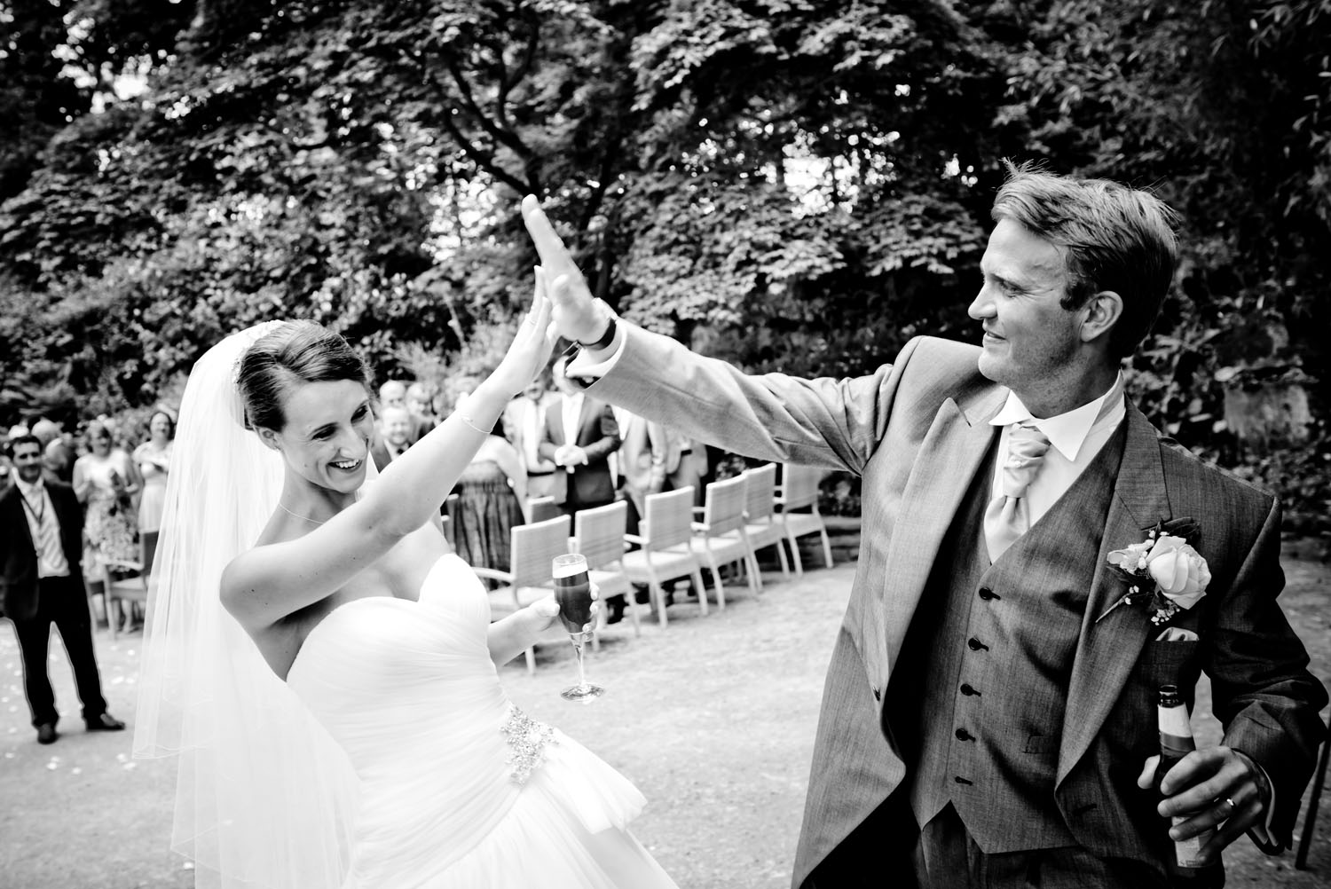Bride and groom celebrating their outdoor wedding with a high five, while guest look on b hind them