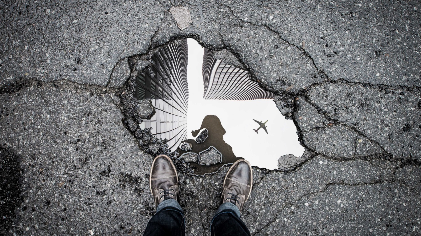 Buraco no asfalto da rua com uma poça de água na qual se vê o reflexo do fotografo.