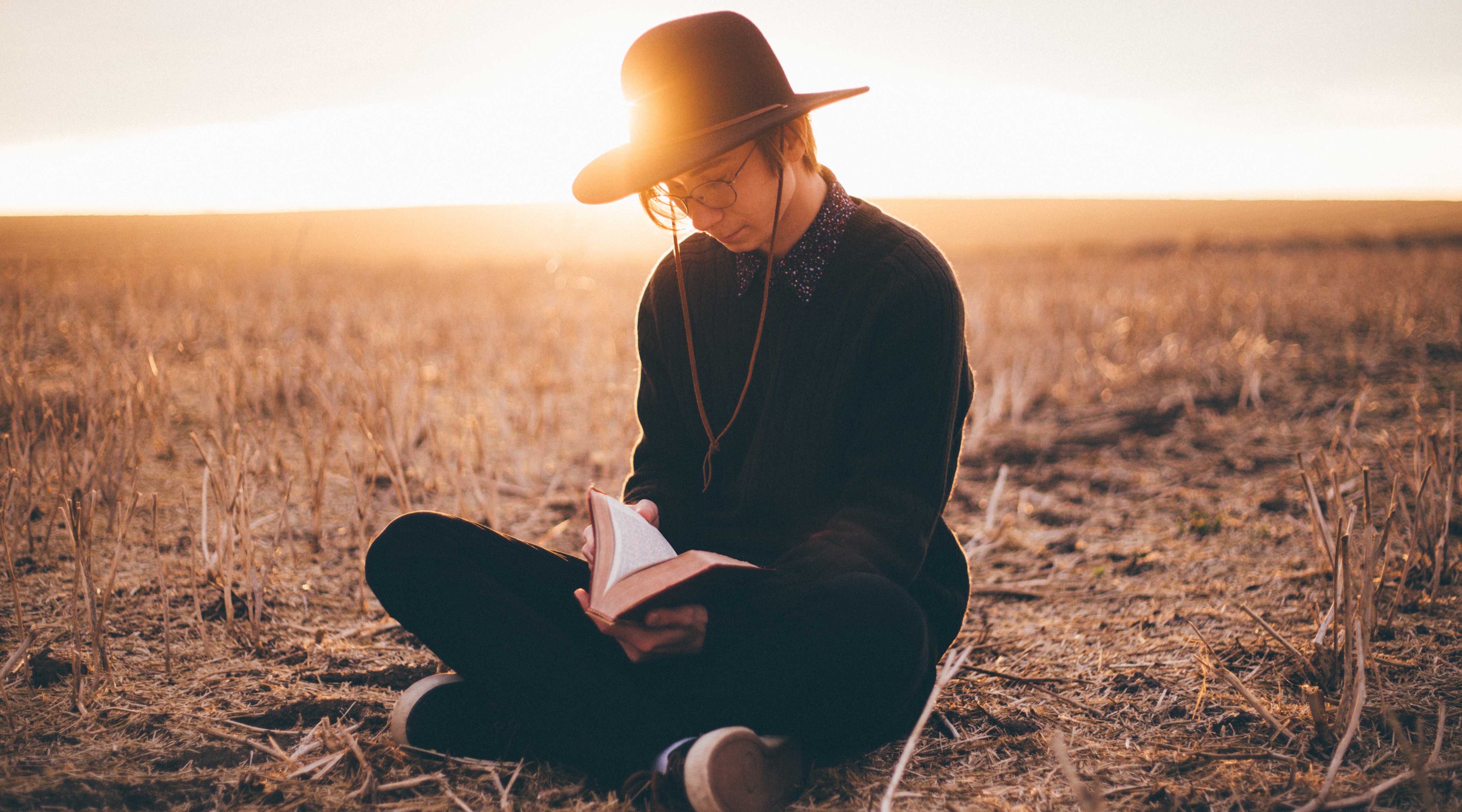Homem sentado no chão de um campo com um livro aberto em seu colo