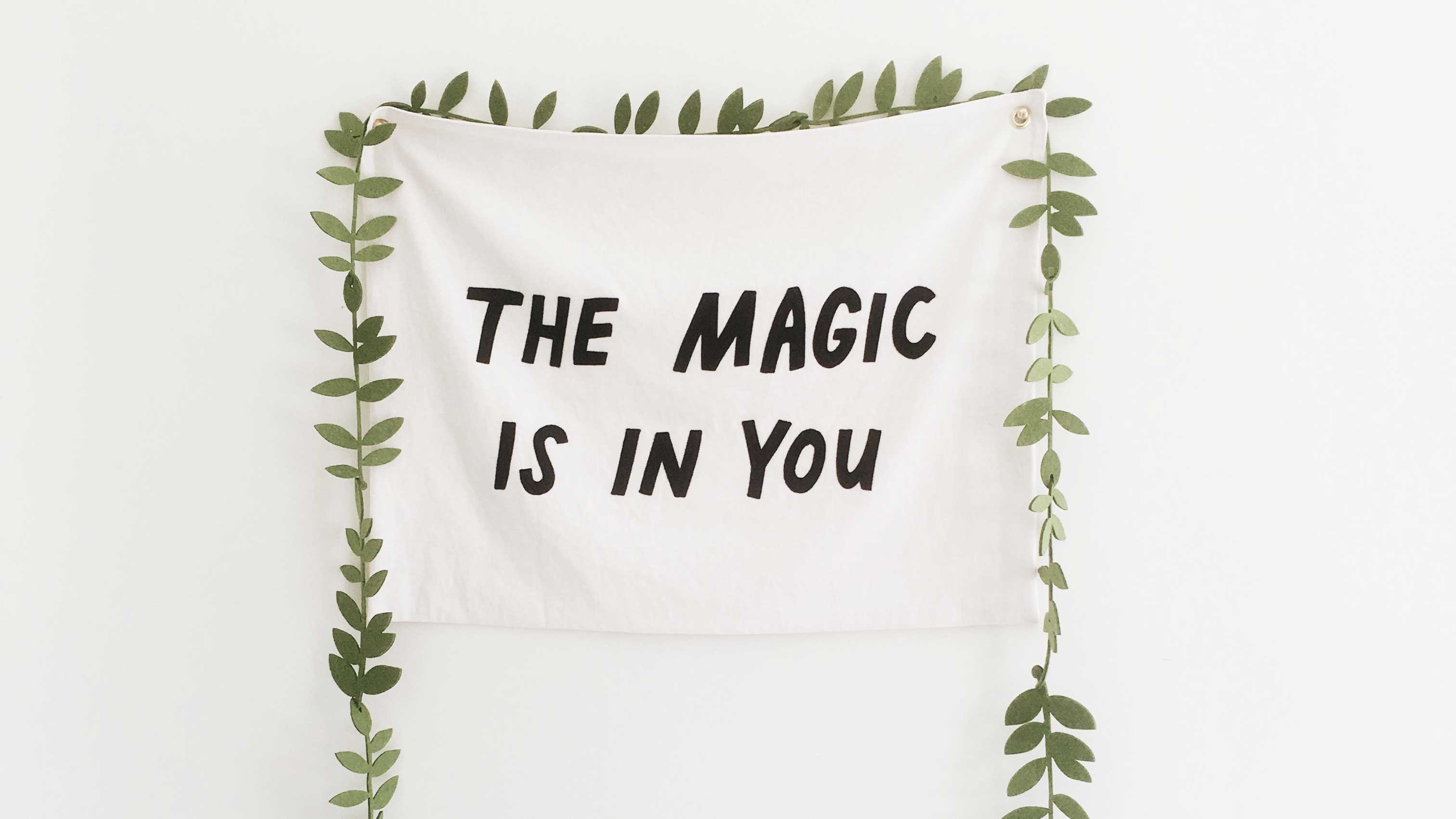 Pano com a frase "Tha Magic is in You" pregado a parede com folhas ao redor, como as de uma árvore.