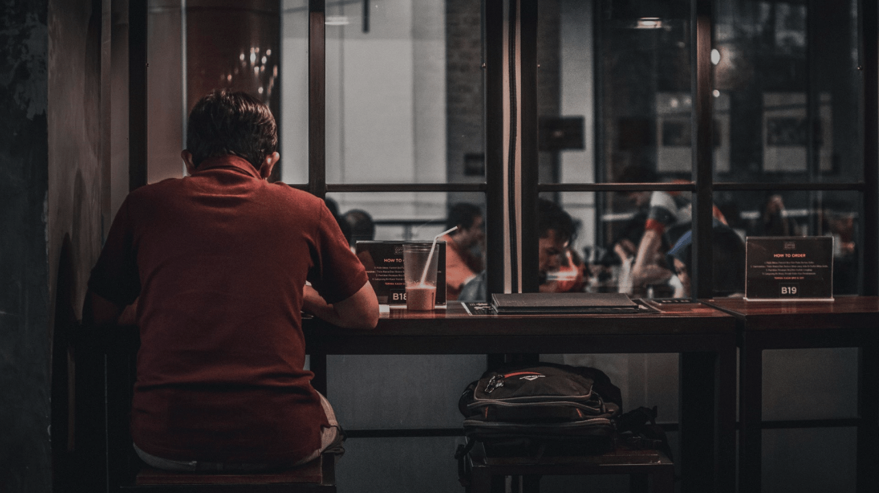 O cenário parece com o de uma cafeteria, temos um homem de costas usando uma camisa vermelha sozinho.