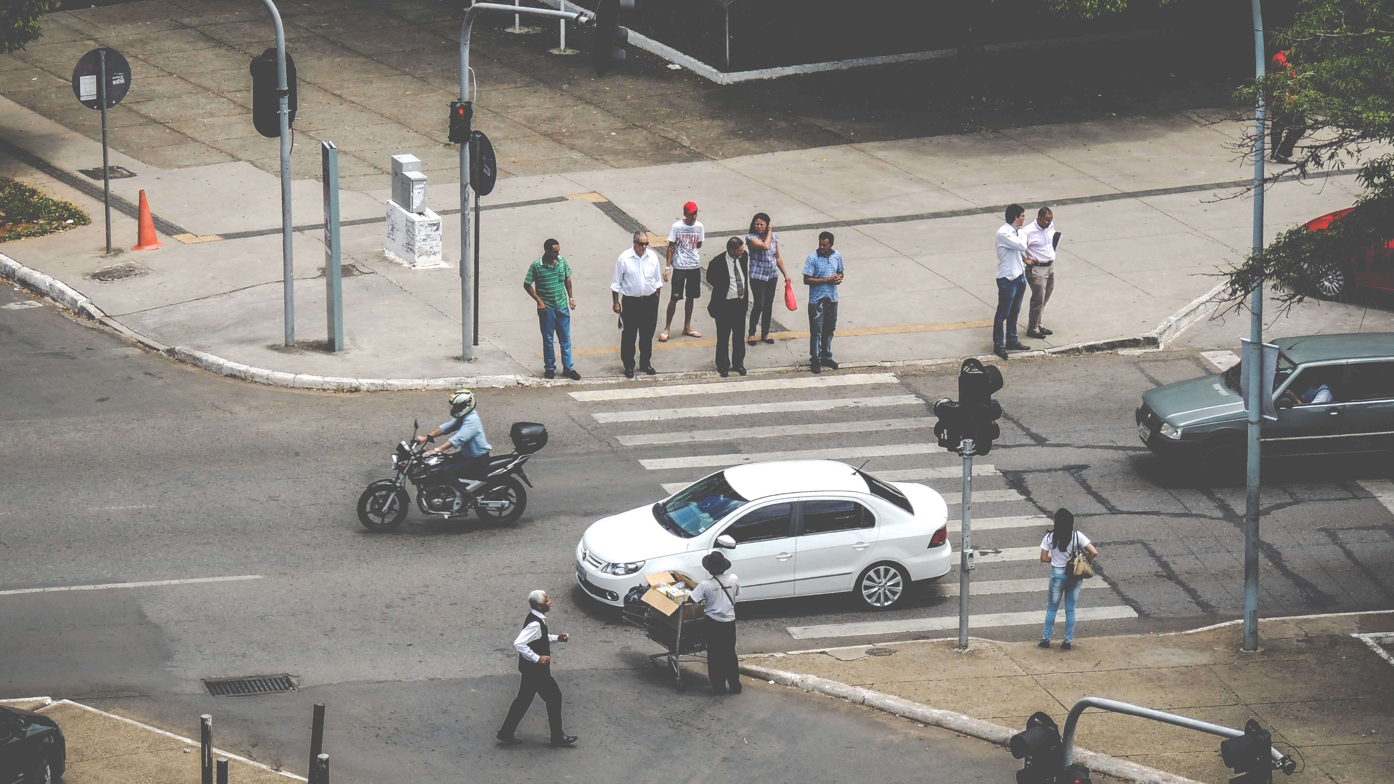 Rua vista de cima com pedestres aguardando para atravessar na faixa enquanto um carro branco passa.