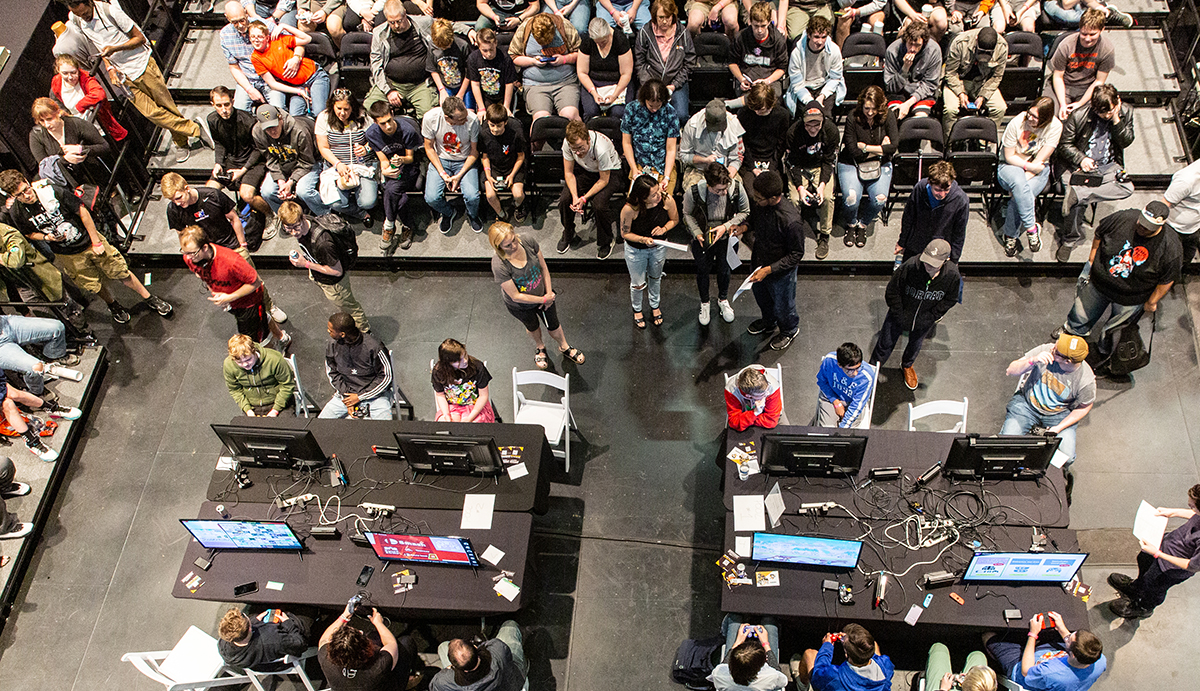 A picture taken from above shows an audience of at least 50 people watching two gaming stations in the middle of the room.