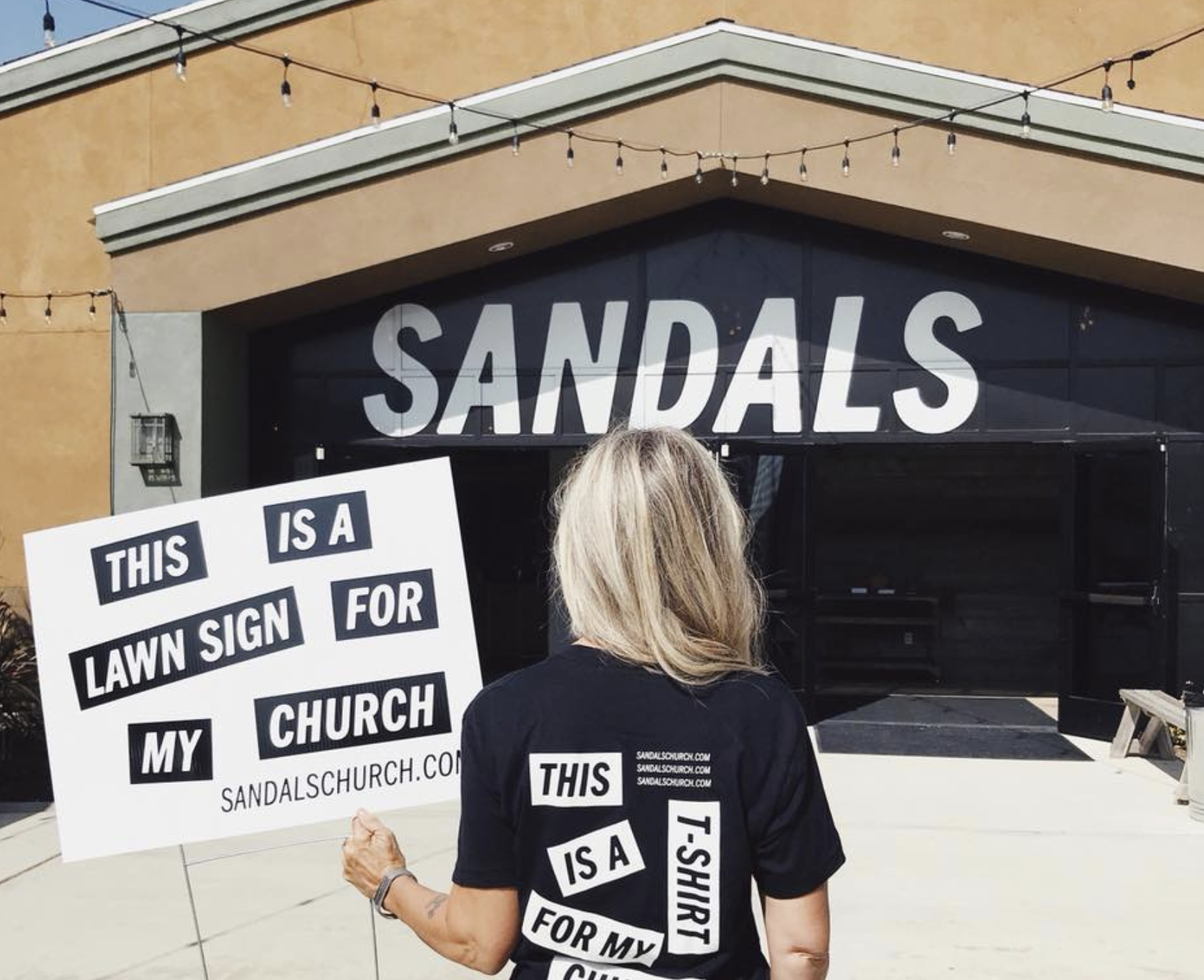 Blonde woman holding a sign that says, "This is a lawn sign for my church" with the url sandalschurch.com at the bottom right.