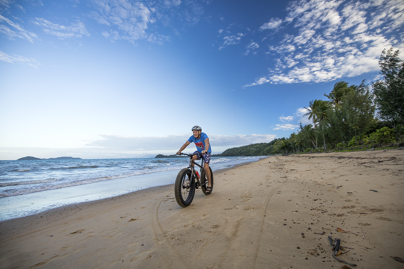 Photo of a man riding a bike along a beach with water and trees in the background.