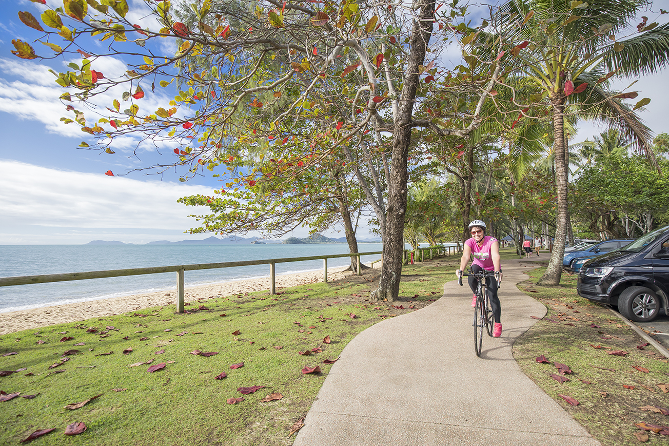 Photo of a woman riding a bike on a bike path with the ocean and trees in the background.