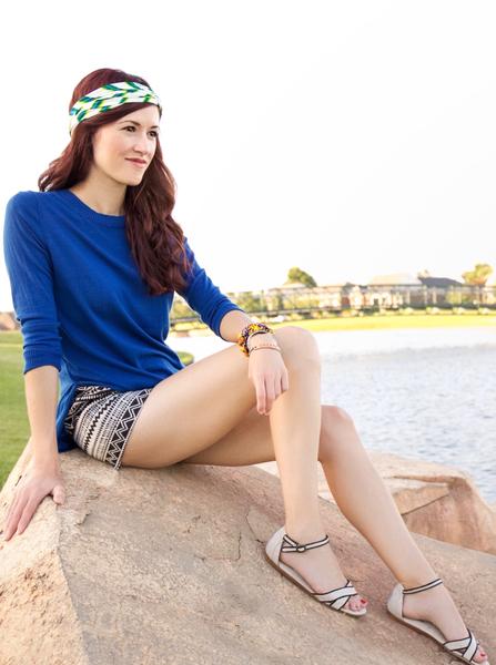 Girl sitting on a rock by a lake. She's wearing TOMs sandals, ivory and black patterned shorts with a deep blue sweater top and Regenerous Designs green big braided headband.