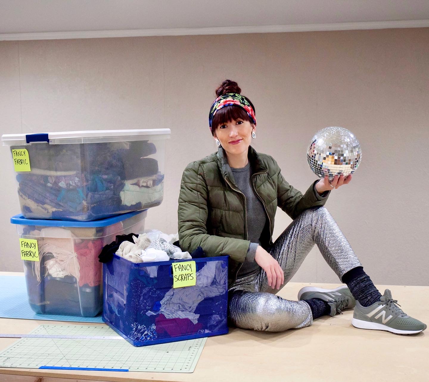 Alyssa Bird wearing Regenerous Designs accessories sitting on a table with boxes of fabric and holding a disco ball
