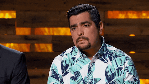 GIF of a man sitting down, tilting his head to the left, and lifting his eyebrows conveying the question "Say that again?"
