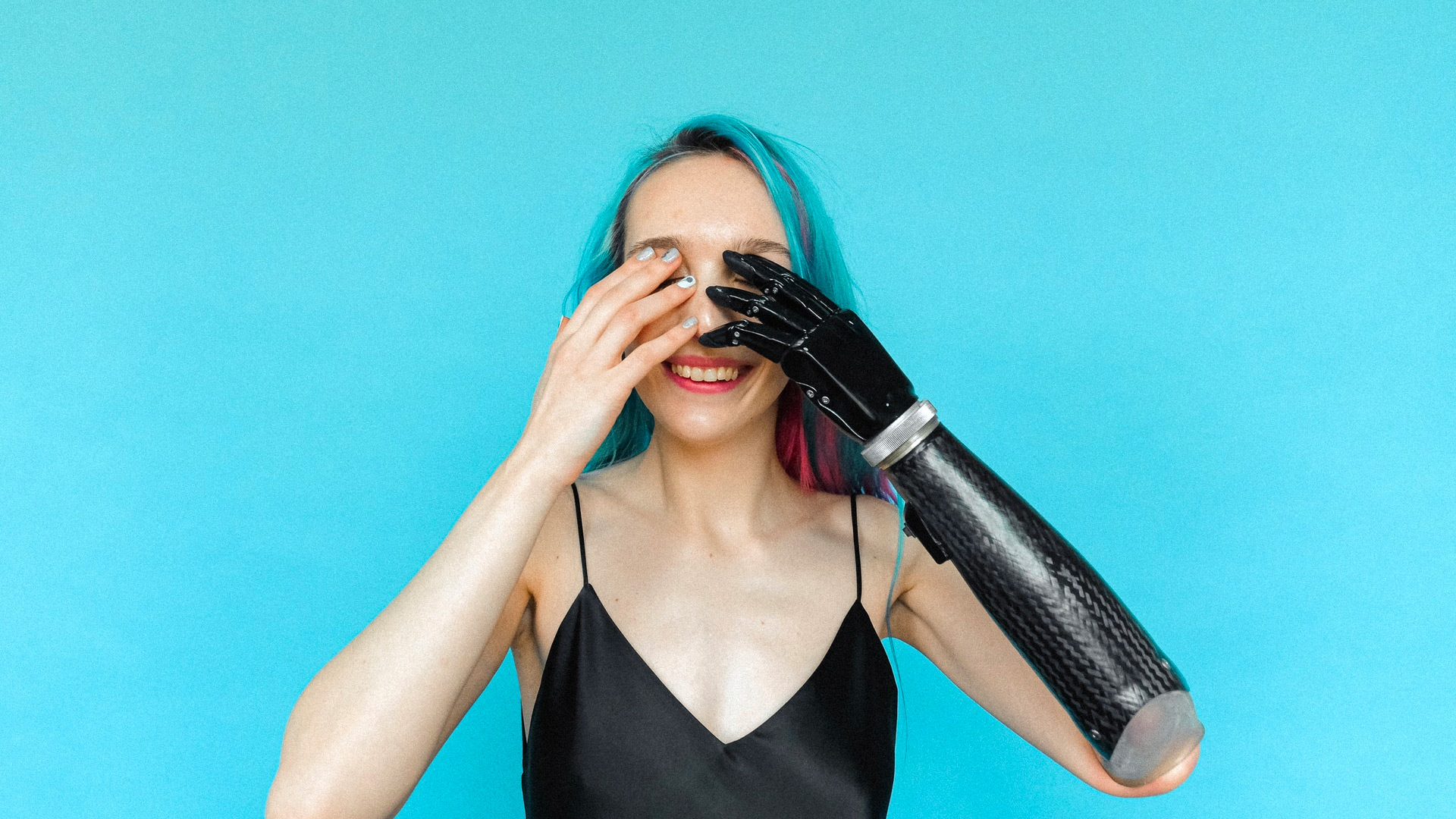 Kvinnan på bilden ler och håller för sina ögon, ena handen är en robotprotes. Hon har ljus hy och färggrannt blått / rosa / lila hår och bär ett svart tunt linne.