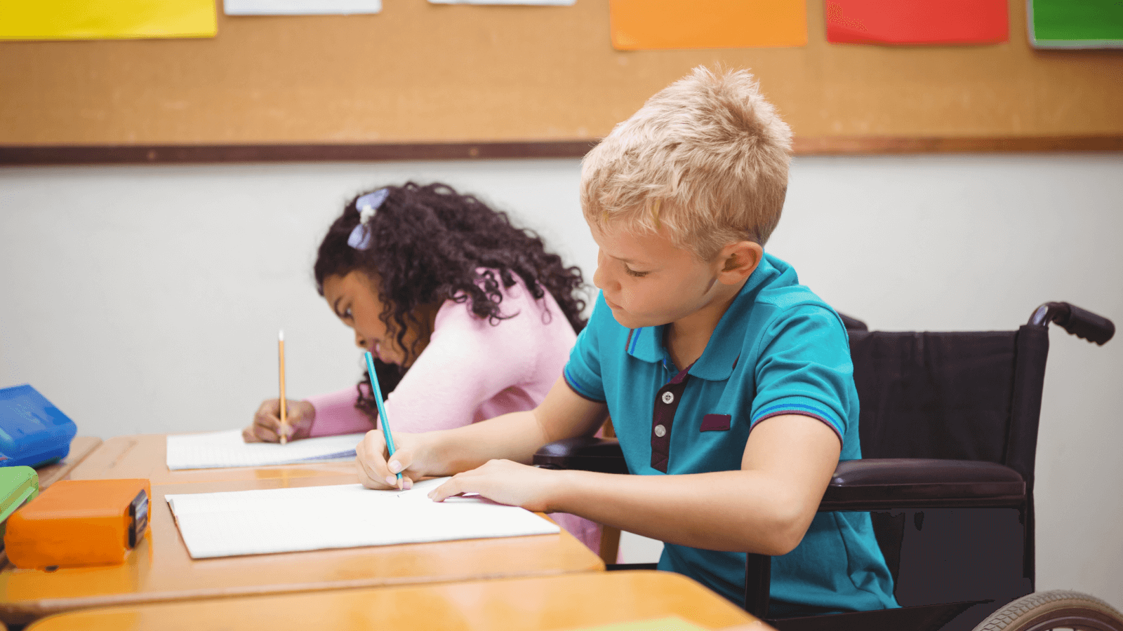 Disabled boy at the table next to other student writing in his book