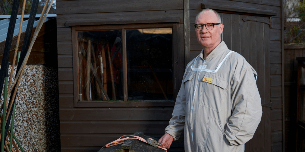 Man in grey boiler suit wearing glasses stands in front o a dark brown shed outside