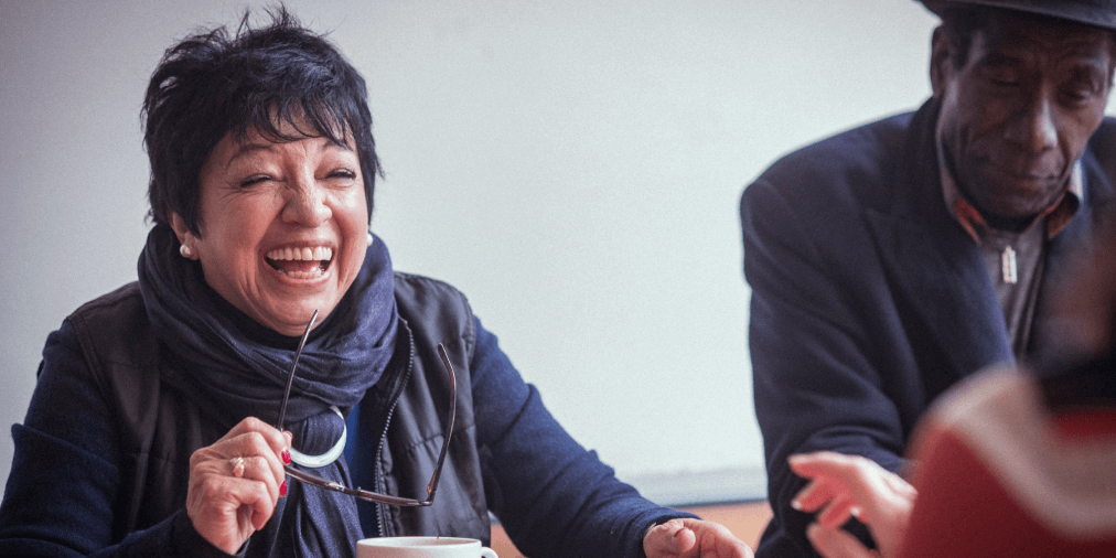 Woman in blue jumper with short black hair and pearl earrings holds her glasses in her left hand while laughing at a person opposite her. A black man in a hat is sitting next to her