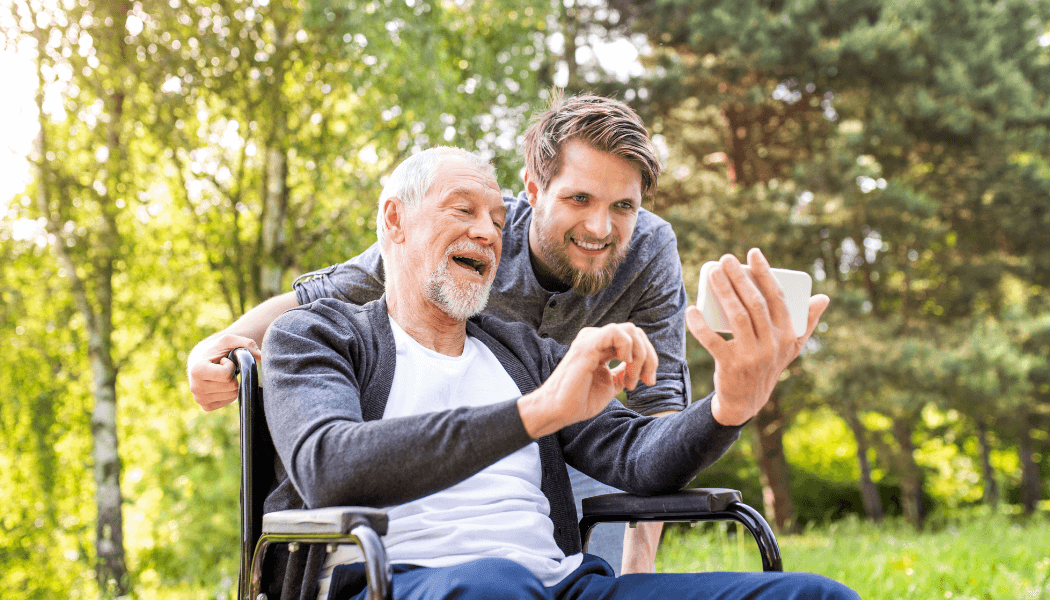 Male carer with beard poses for a photo with an elderly man in a wheelchair who is holding a white phone in his outstretched hand and smiling.