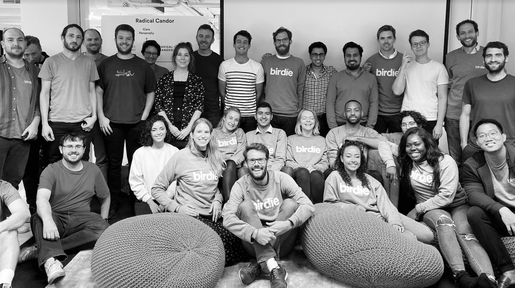 Team of people stand in office, in front of radical candor poster, wearing grey birdie jumpers. There are beanbags in the forefront