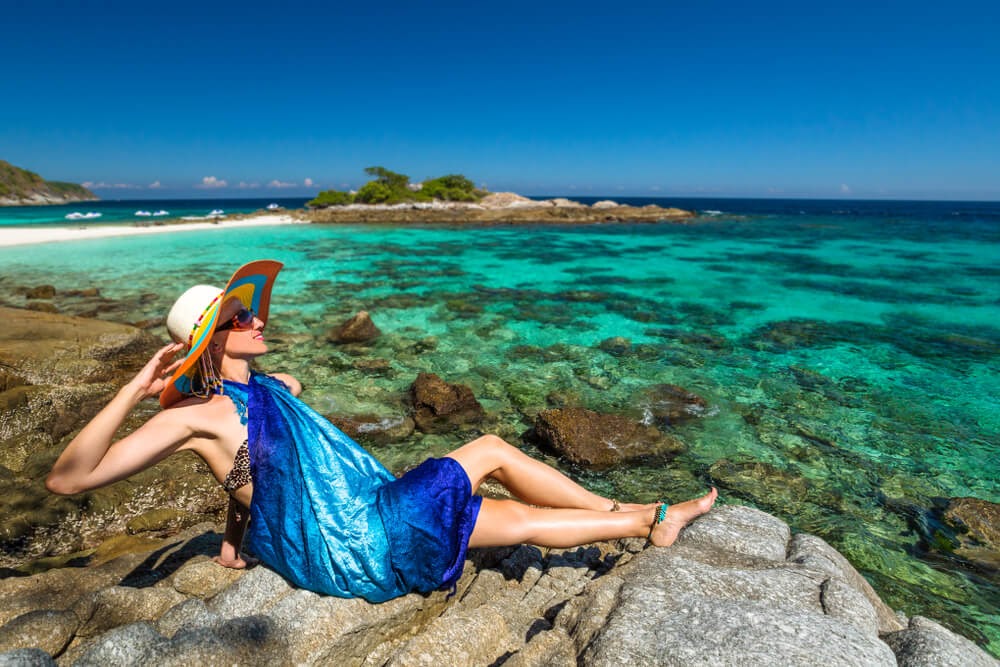 Woman in sarong and sunhat lay on beach with beautiful clear blue water surrounding her.