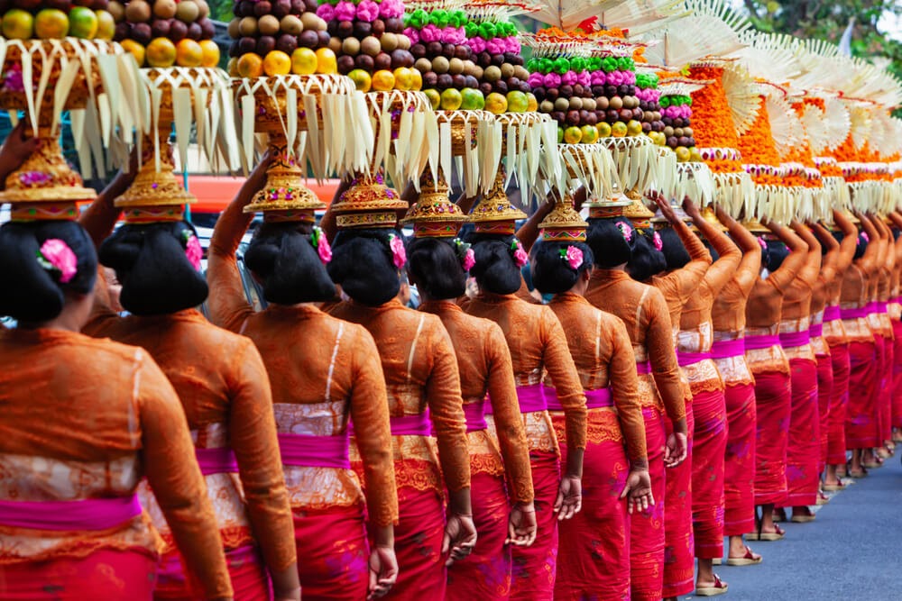 Photo of a line of women from behind in brightly colored clothes and sarongs. They are holding elaborate headpieces with various fruits and nuts