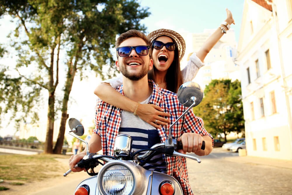 Young couple ride on small motor cycle. Woman holds onto man from behind and smiles with one of her arms in the air.