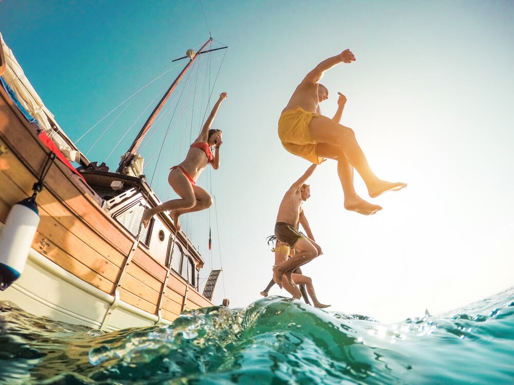 Group of yound people jumping into the ocean from a large sailboat. Photo is taken from below and the sun is shining into the camera.