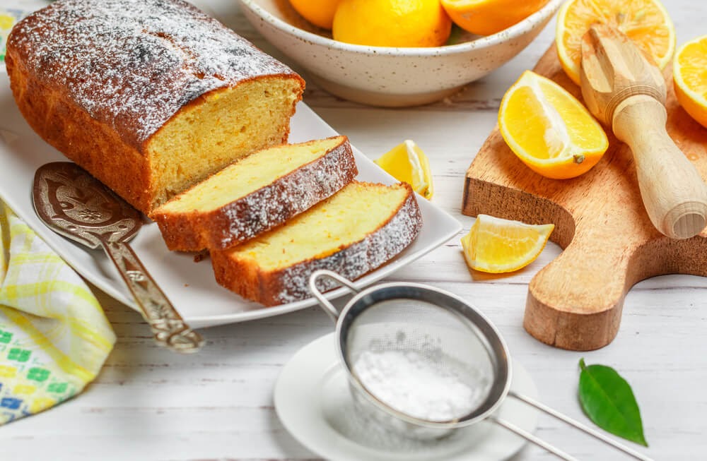 Sliced loaf of lemon bread dusted with powered sugar. A bowl of lemons and s few lemon wedges sit next to the loaf.