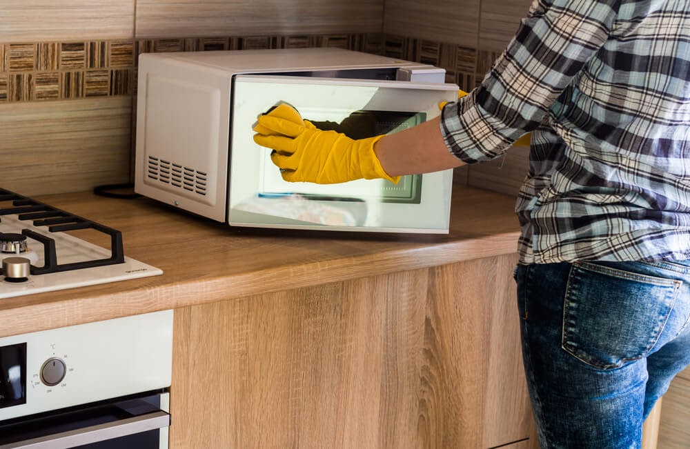 Person in a blue plaid shirt and jeans scrubbing their microwave in yellow latex glovese