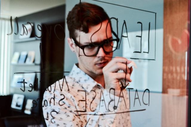 man with glasses writing on a glass board