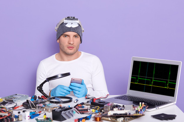 Young computer specialist repairing broken smart phone, being ready to dismantle it, siiting at table full of tools, radiotrician testing electronic equipment in service centre. electronic engineering Free Photo