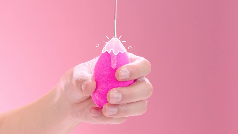 Hand holding a beauty blender sponge on a pink background
