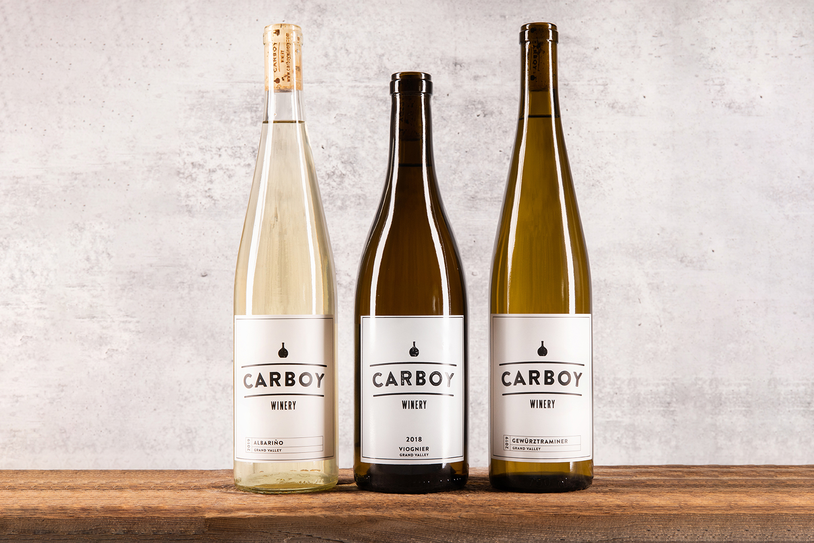 Three bottles of Carboy Winery wines sit on a wooden table with a concrete background.