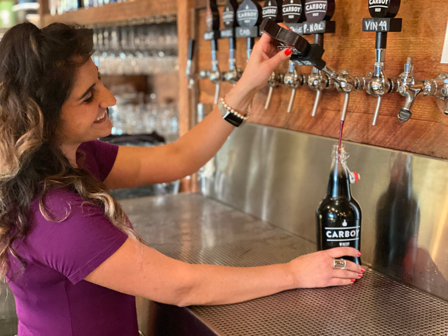 A woman pours smiles and pours wine into a Carboy wine growler from wine taps on the wall at a bar.