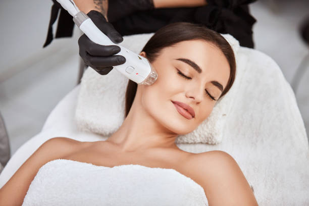 beautiful girl lying in spa salon covering breast with white towel and getting massage for her cheek with professional apparat laser hair removal stock pictures, royalty-free photos & images
