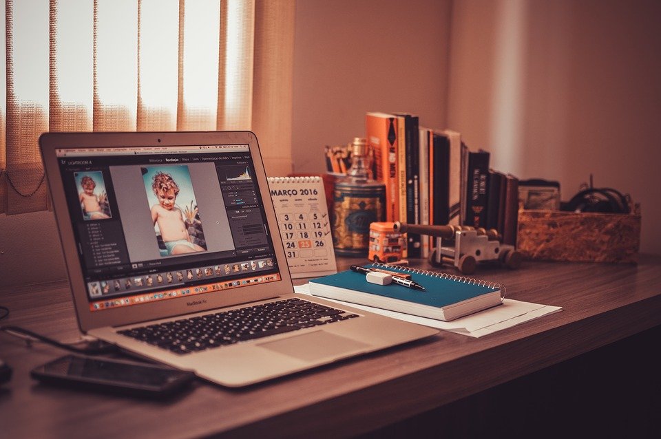 Working from home can prove to be beneficial or detrimental depending on the employee. The picture shows a Macbook, a phone, stationery and a calendar on a wooden workdesk.