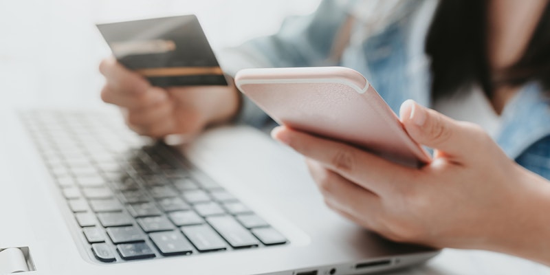 A person holding a smartphone and credit card in front of a laptop