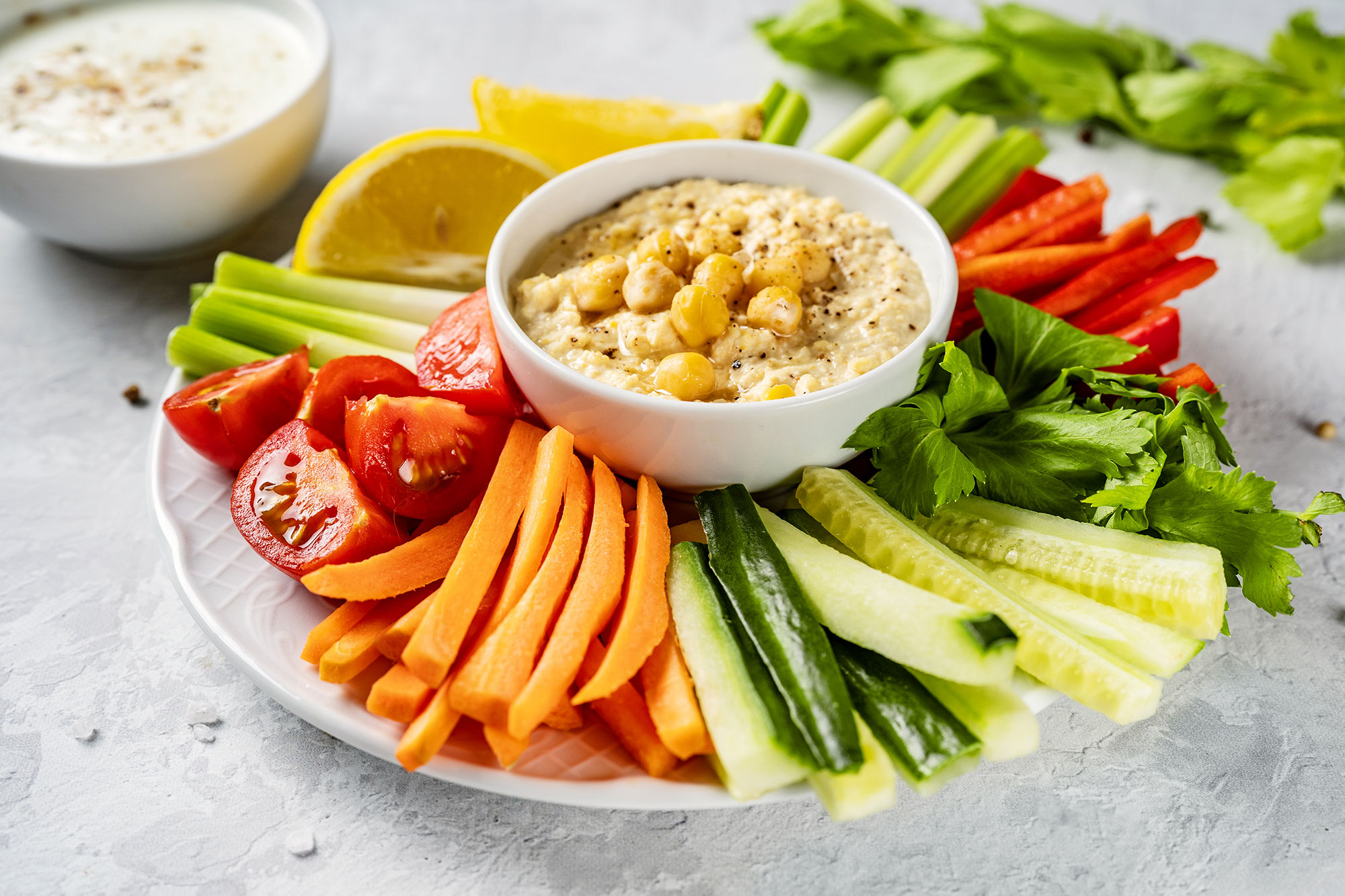 A plate with a bowl of hummus with carrots, cucumbers and tomatoes around it
