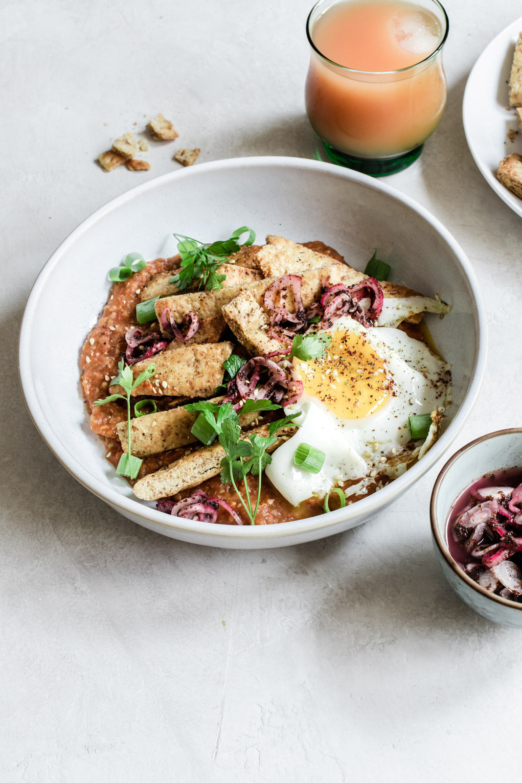 Bowl of eggplant pitaquiles topped with a fried egg and sumac onions with a glass of grapefruit juice