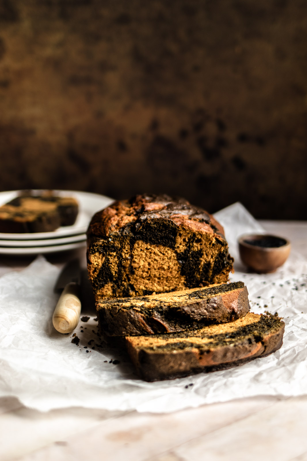 A loaf of black sesame swirl pumpkin bread with 2 slices taken off, a wooden knife and a small wooden pinch bowl