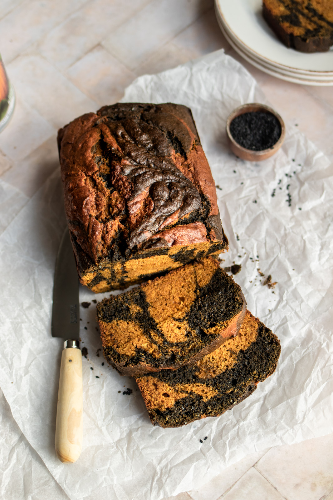 A loaf of black sesame swirl pumpkin bread sliced open and sitting on baking paper with a wooden knife and a bowl of black sesame seeds
