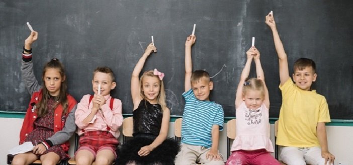 school children sitting in front of a blackboard, holding chalk above their heads