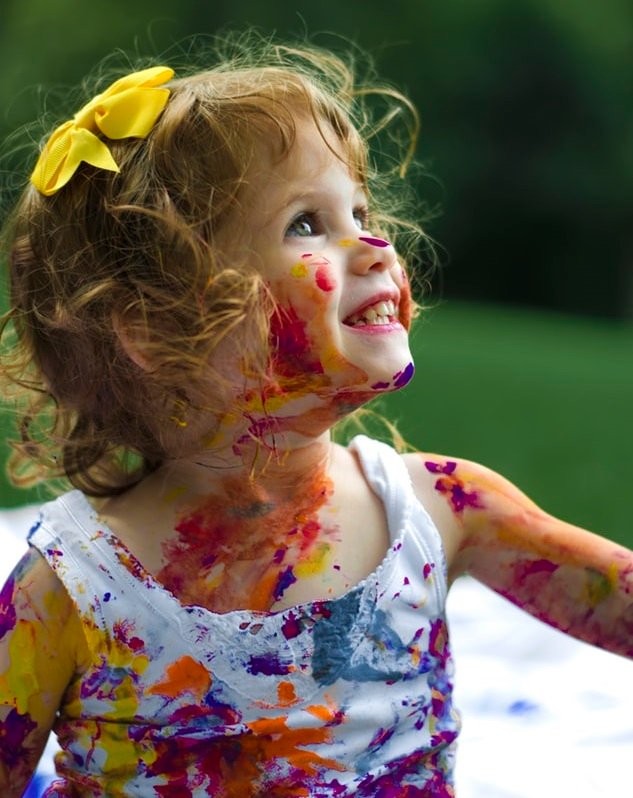 blonde toddler girl covered in messy paint on clothes and face.