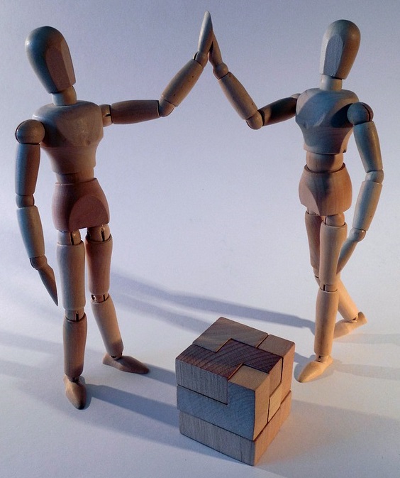 two wooden articulated puppets high-fiving over completed wooden block puzzle cube.