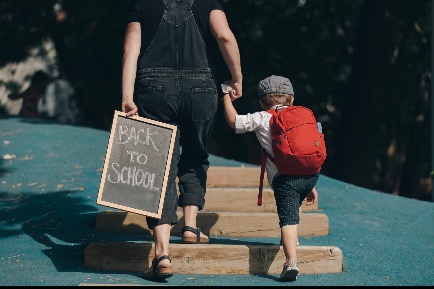 rear view of adult holding a Back To School chalkboard and the hand of a child wearing red backpack.