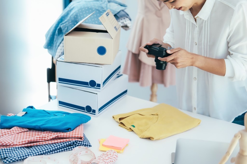 A woman takes photo of several clothing products next to shipping boxes.