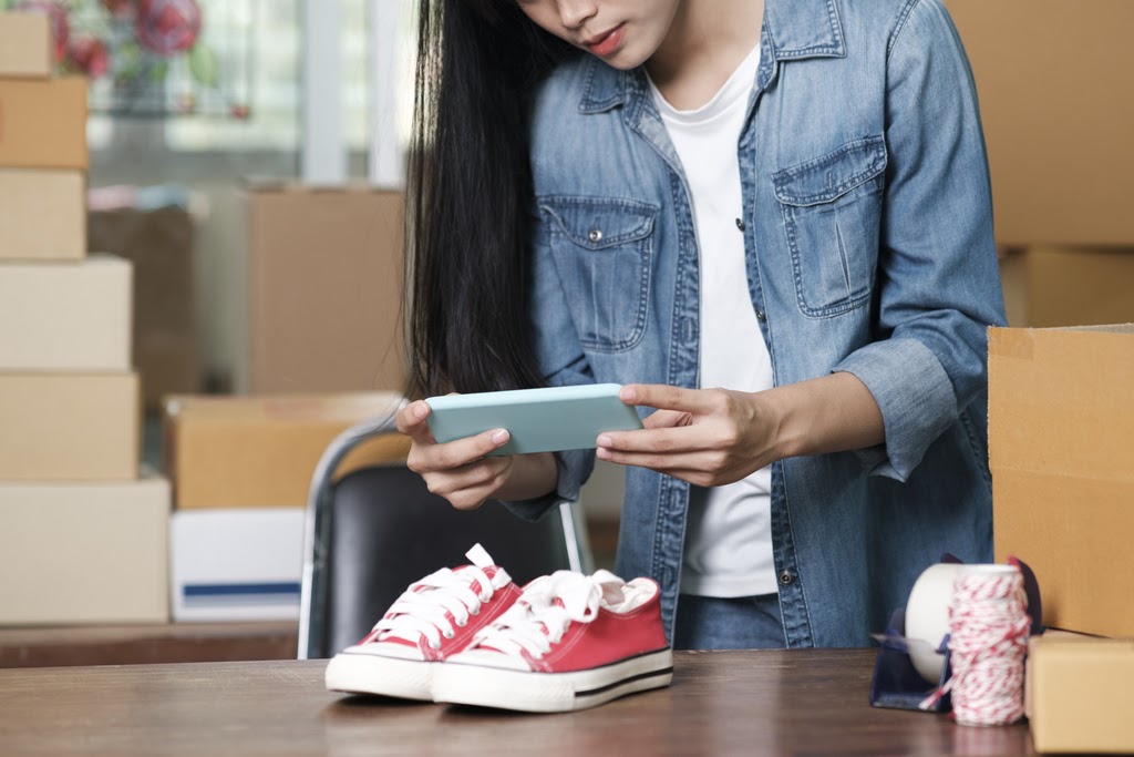 A woman takes a photo of pink shoes with a cell phone next to shipping supplies.