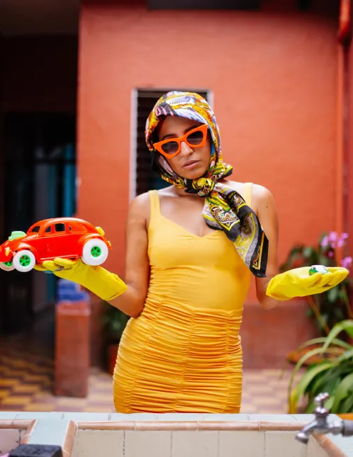 La Lulu standing in front of house wearing cleaning gloves and head scarf holding toy car and diamond.