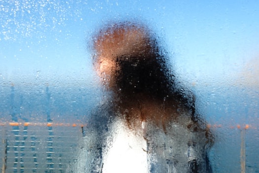 A blurred view of a white woman through a window, wet and obscured. She wears a white shirt and blue jacket, has long brown hair, and is looking to her right. Vast blue sea and sky is behind her.
