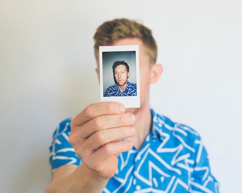 A white man with brown hair wearing a graphic-printed blue and white short sleeved button up holds a small polaroid photo in front of his face. On the polaroid, we see the same man in the same shirt, his gaze averted slightly from the camera.