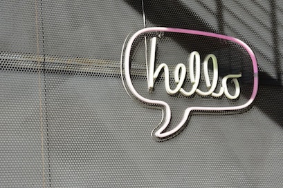 on a textured grey grated-metal wall, a white neon speech bubble with the word "hello" in neon cursive letters