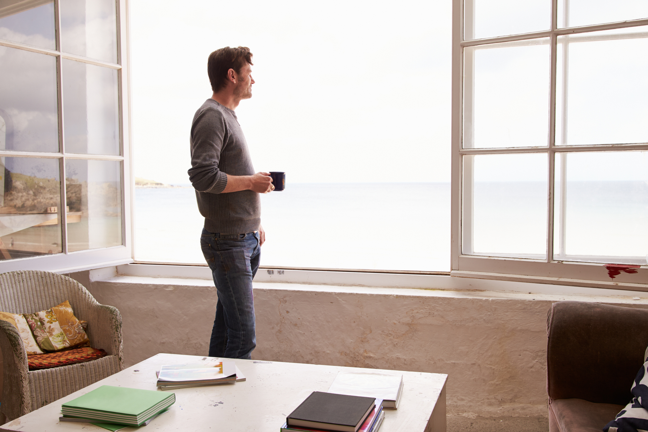 A man with a cup of coffee staring at the ocean through his window