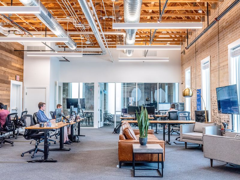 Grand open space style industriel avec des gens qui travaillent espacés sur des bureaux en bois et métal placés devant des canapés, tables d'appoint avec plantes et fauteuils.