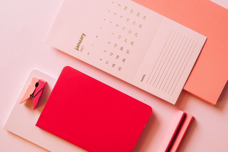 Un calendrier blanc de janvier 2021 posé sur un fond rose à côté d'un carnet rouge et d'une pince à linge rose